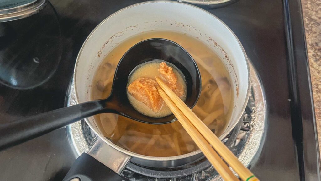 miso and a small amount of dashi in a ladle, chopsticks mixing the liquid in the ladle over the miso soup in a pot.