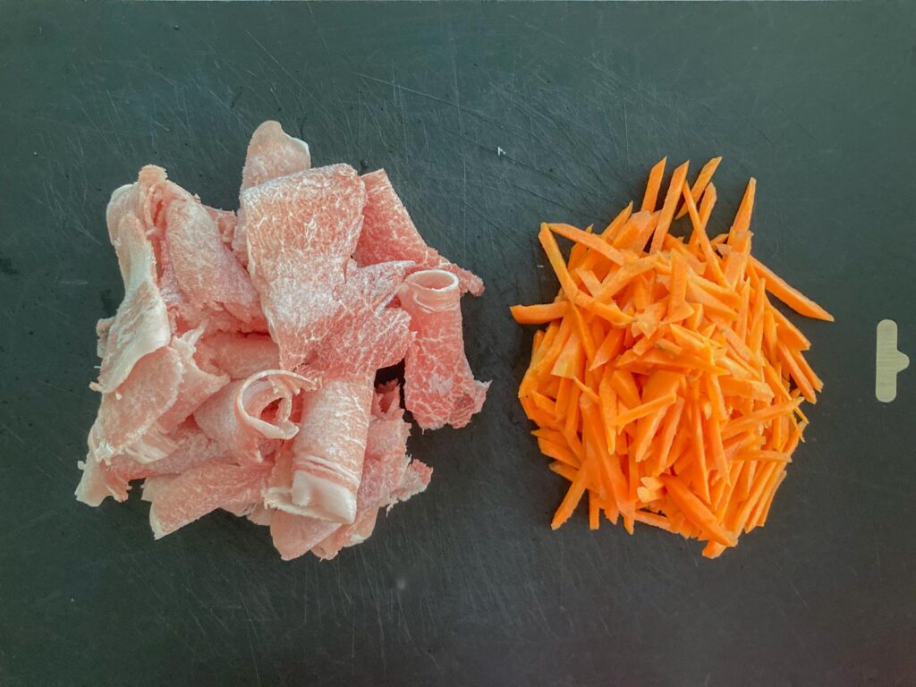 sliced pork on the left and julienned carrots on the right on a black cutting board