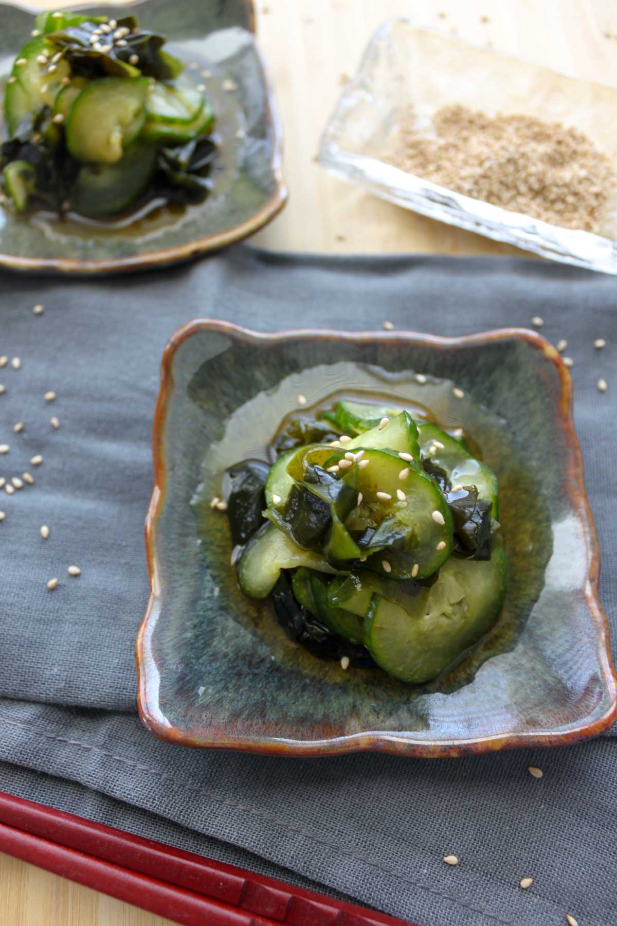 two square plates with sunomono (Japanese cucumber salad) salad topped with white sesame seeds.