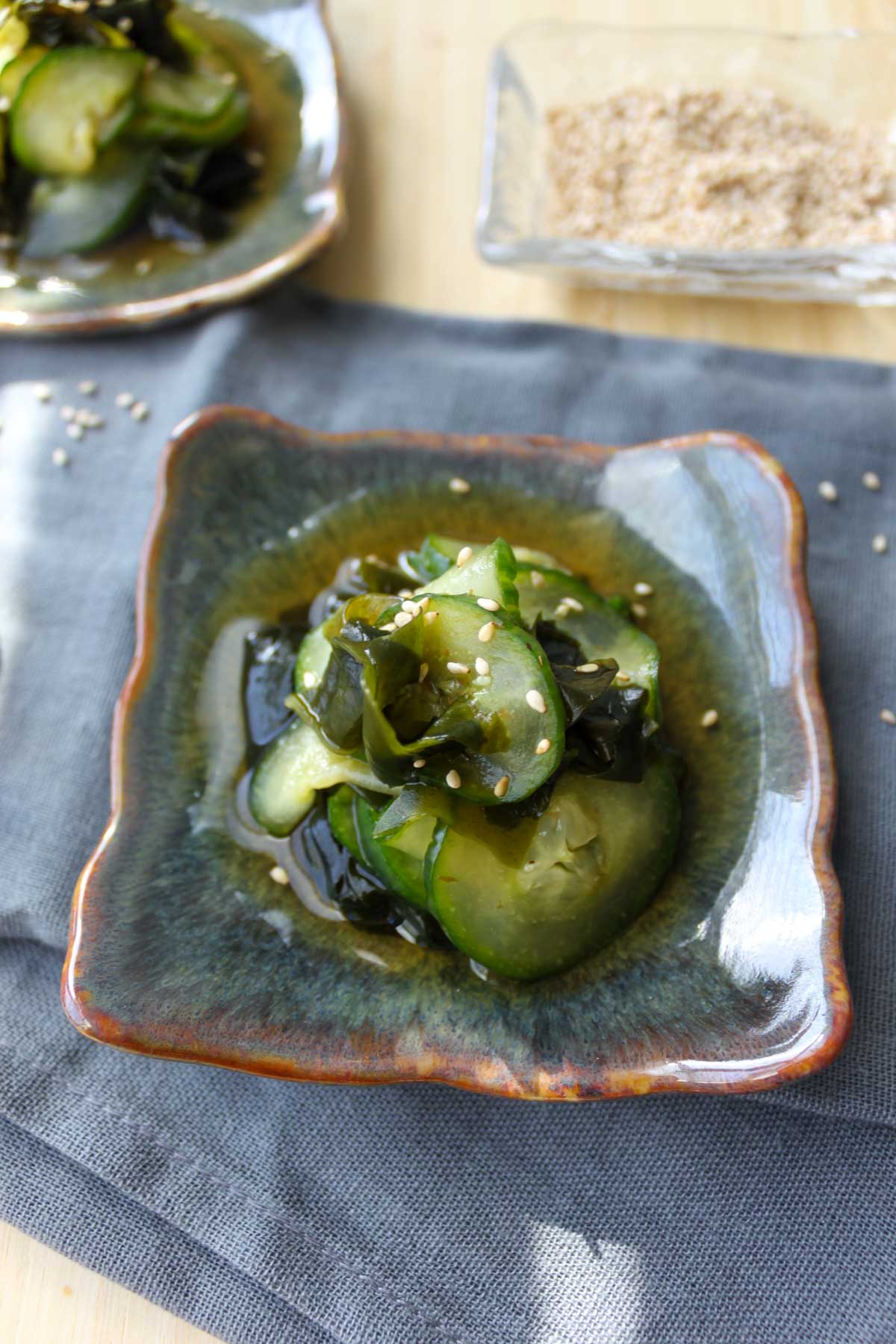 two square plates with sunomono (Japanese cucumber salad) salad topped with white sesame seeds.