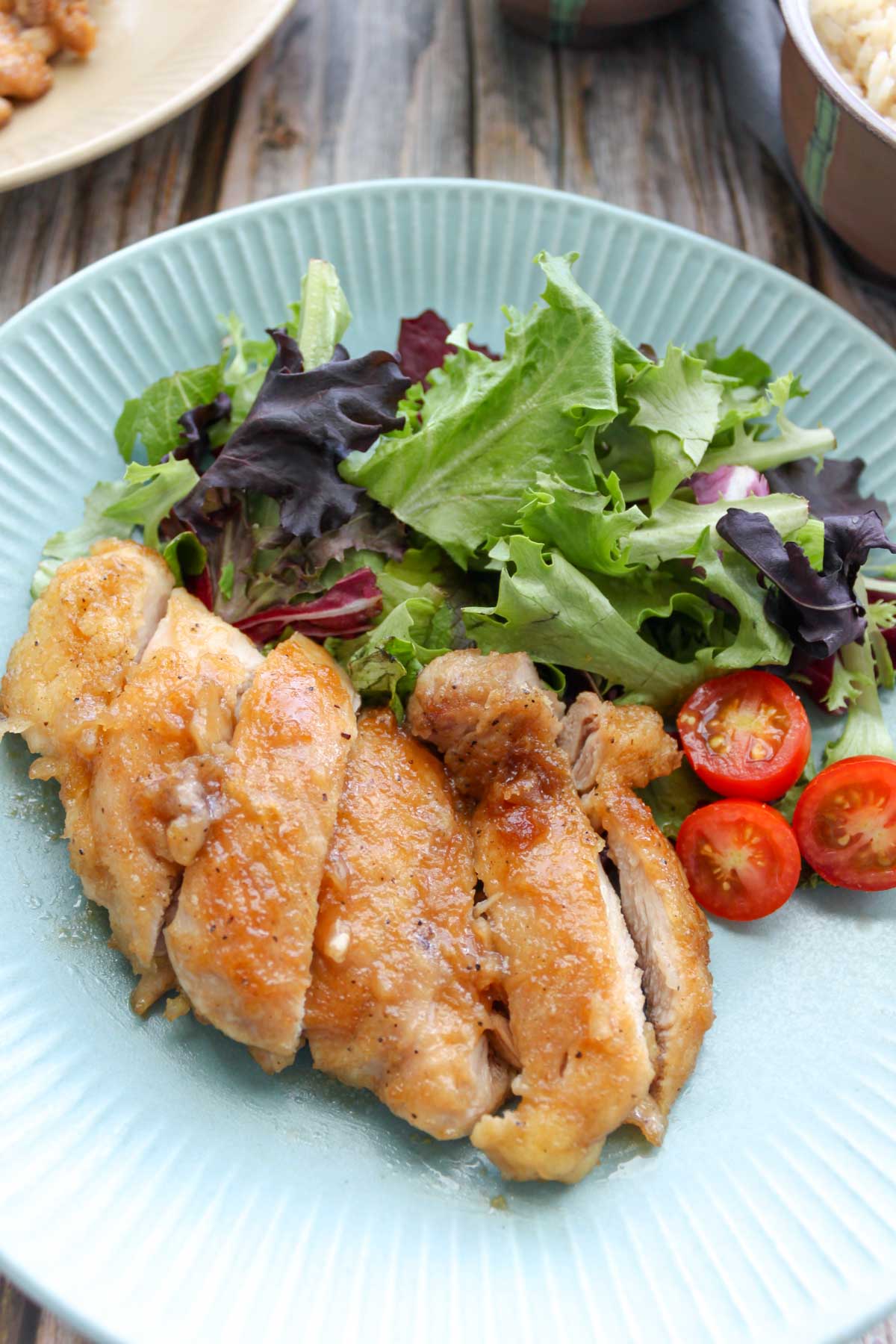 a plate with green mix, cherry tomatoes, and sliced teriyaki chicken. there is a bowl of rice at the top right corner.