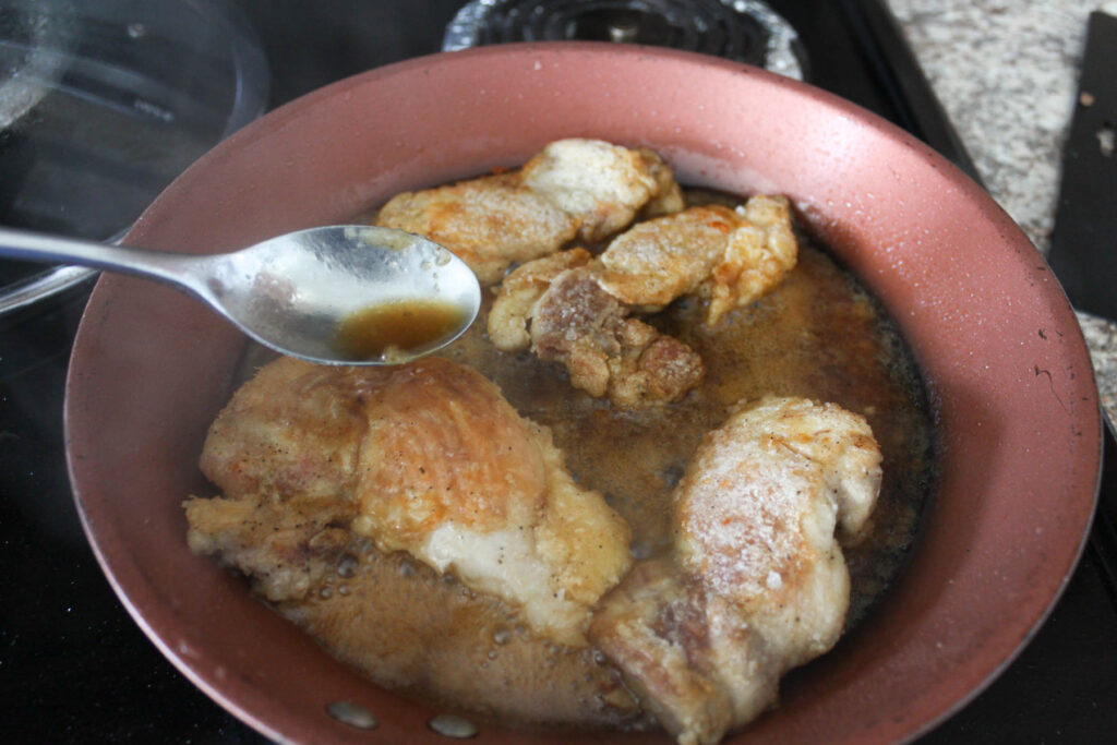 four chicken thigs and teriyaki sauce on a frying pan, and a spoon is pouring the sauce over the chicken.