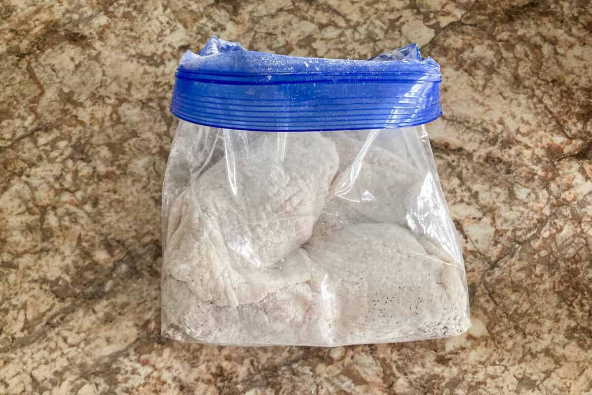 Two pieces of chicken thighs are coated with cornstarch in a plastic bag.