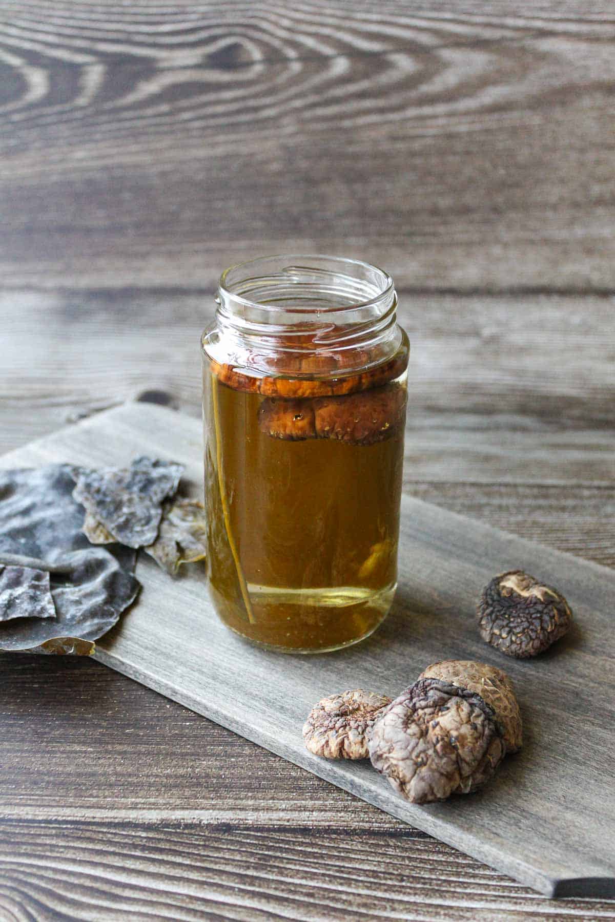 Vegan dashi (kombu and dried shiitake mushrooms) in a glass bin, surrounded by dried shiitake and kombu on the side.